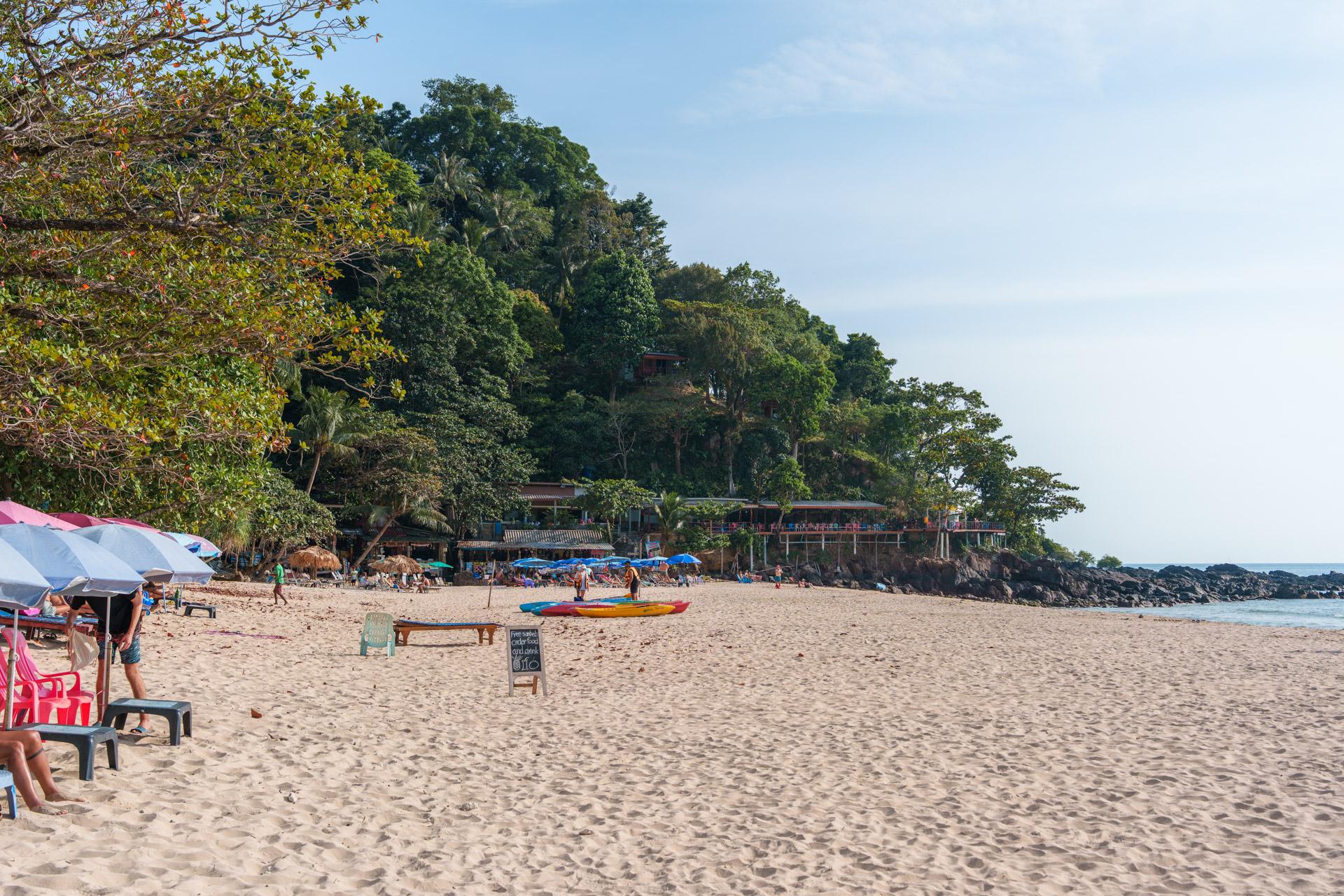Charlie Beach on Koh Mook, with sandy shore, colourful umbrellas, kayaks, dense tropical vegetation and seaside structures.