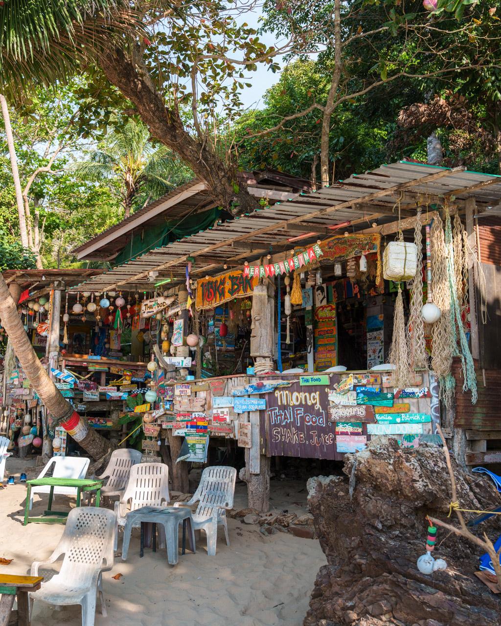 Rustic beachfront Thai food restaurant with wooden shack, colourful signs, plastic chairs and hanging decorations among palm trees.