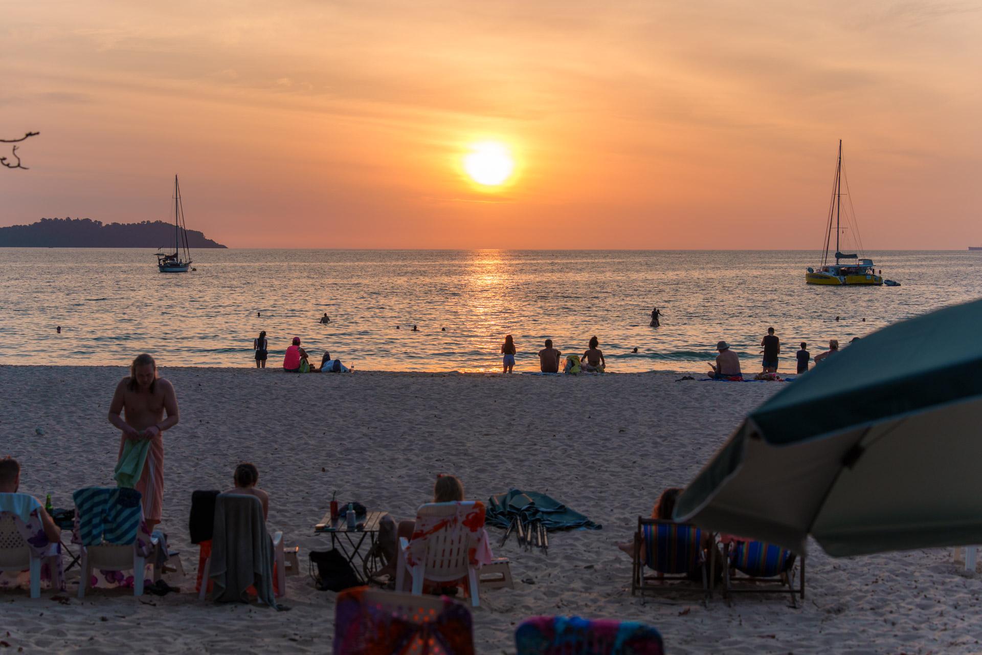 Sunset beach scene at Charlie Beach on Koh Mook, Thailand, with swimmers, sailboats, beachgoers and umbrellas along the shore.