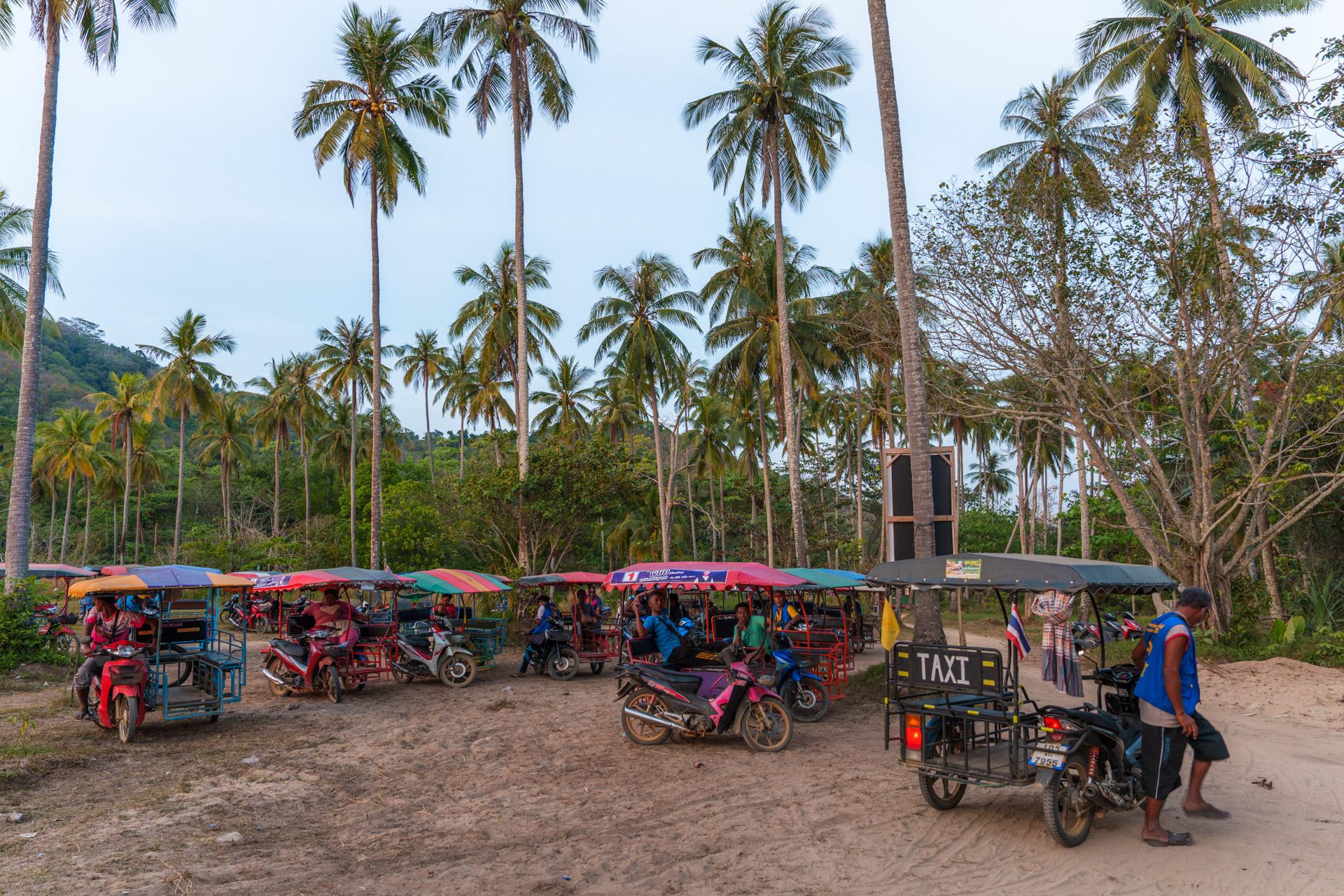 Colourful tuk-tuk taxis lined up on sandy ground with tall palm trees and forested hills at Koh Mook, Thailand.