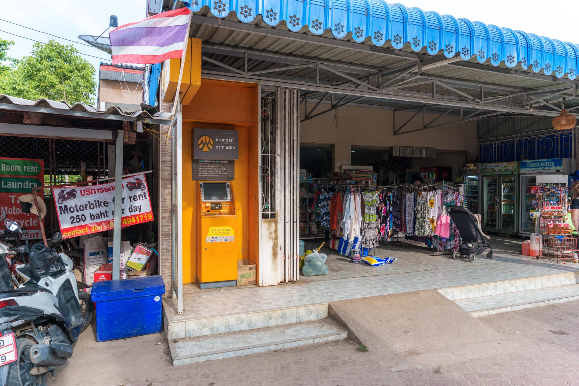 Koh Mook Supermart with Thai flag, ATM, motorbike rentals and colourful clothing displayed outside.