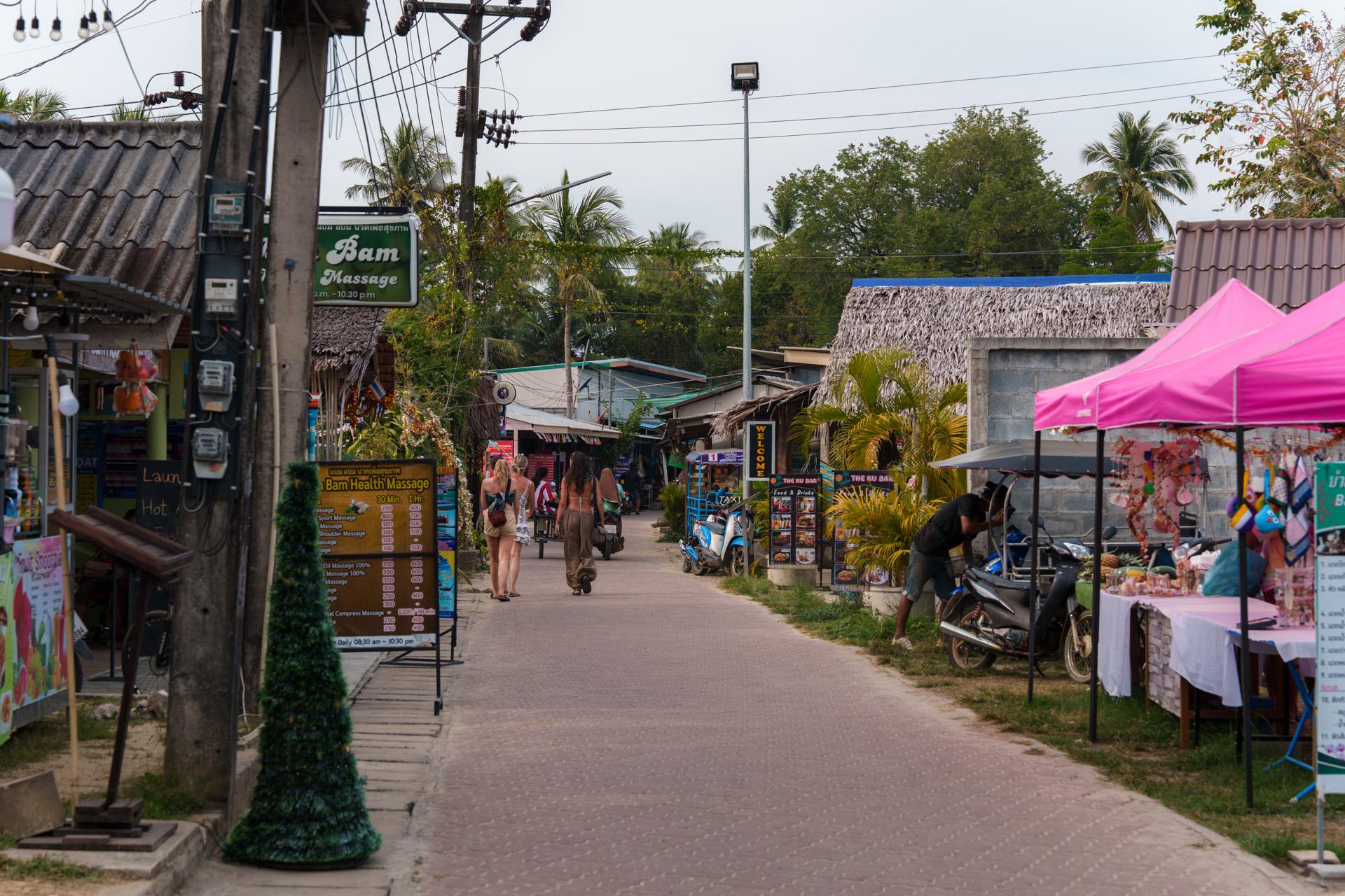 Koh Mook village street with massage shops, thatched-roof buildings, a pink vendor tent, palm trees and tourists walking.