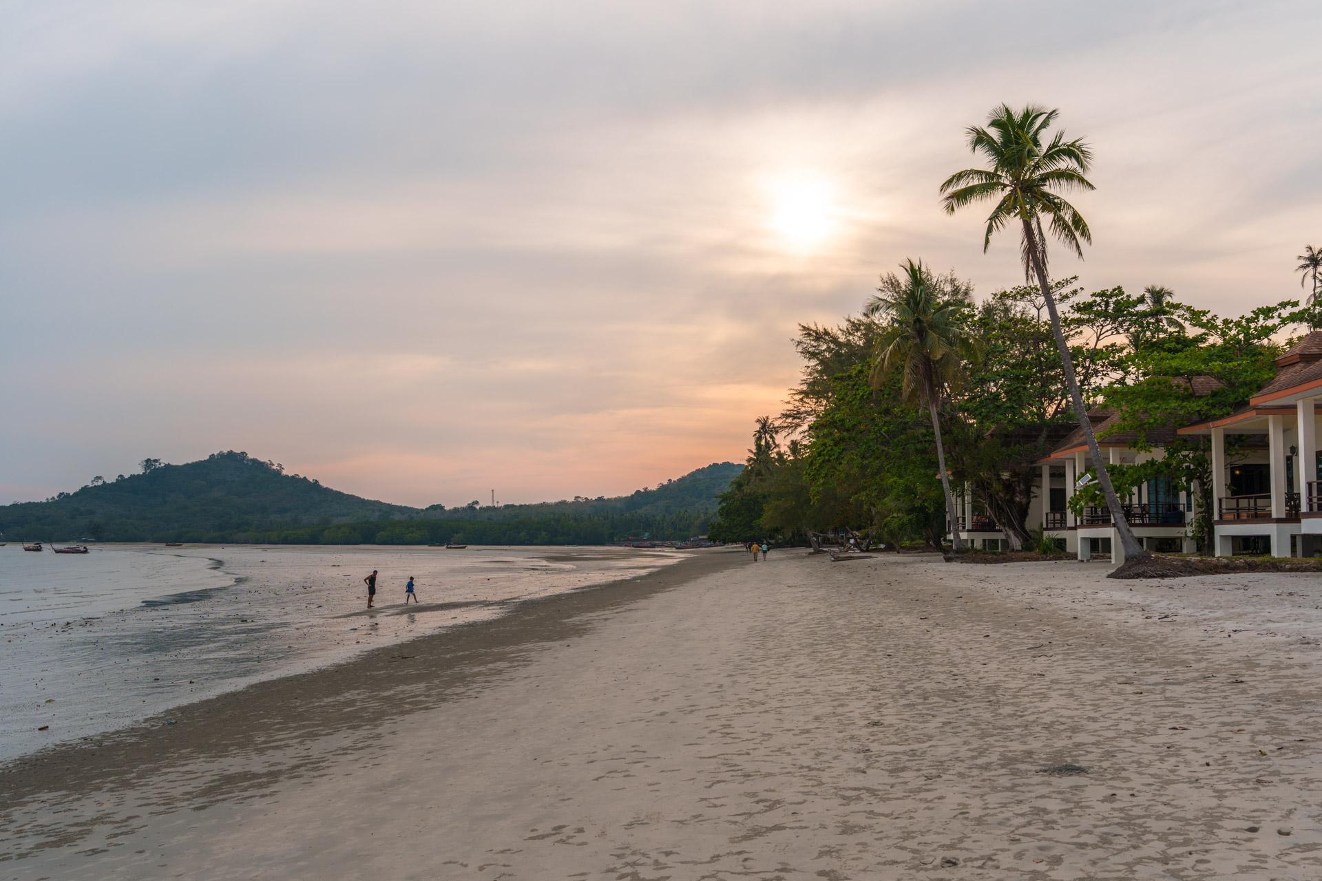 Koh Mook beach at sunset with sandy shore, palm trees, white beachfront building and forested hills across calm waters.