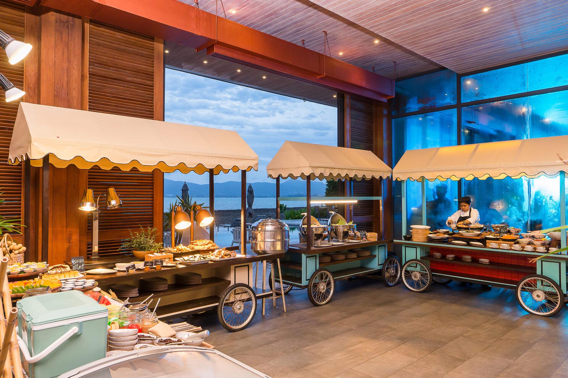 Multiple vintage-style food carts with cream awnings and wheels displaying Thai dishes at beachfront resort dining area at dusk.