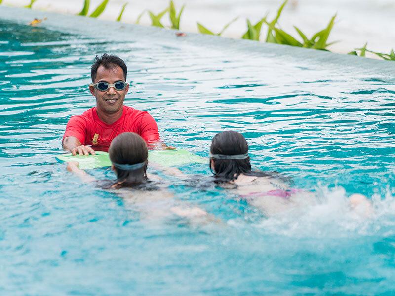Adult instructor in red shirt and sunglasses teaches two children swimming in tropical resort pool with green plants visible.