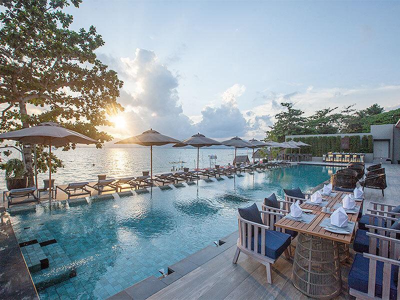 Luxurious beachfront pool at Phuket resort with loungers, umbrellas, dining area, and ocean sunset view.