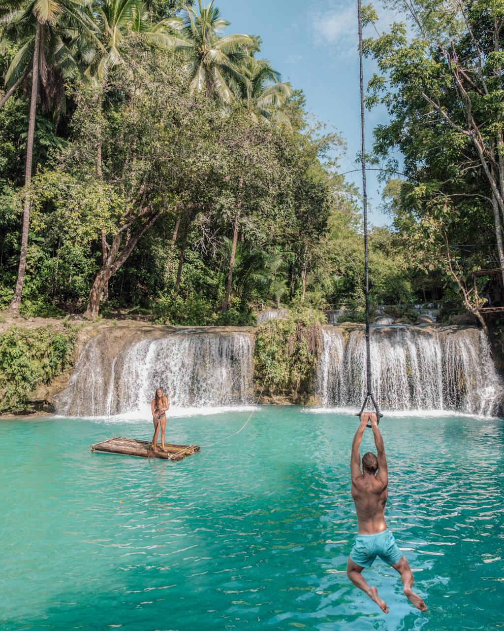 Alex swings from a rope towards Victoria in turquoise water beneath a waterfall on Siquijor, Philippines.