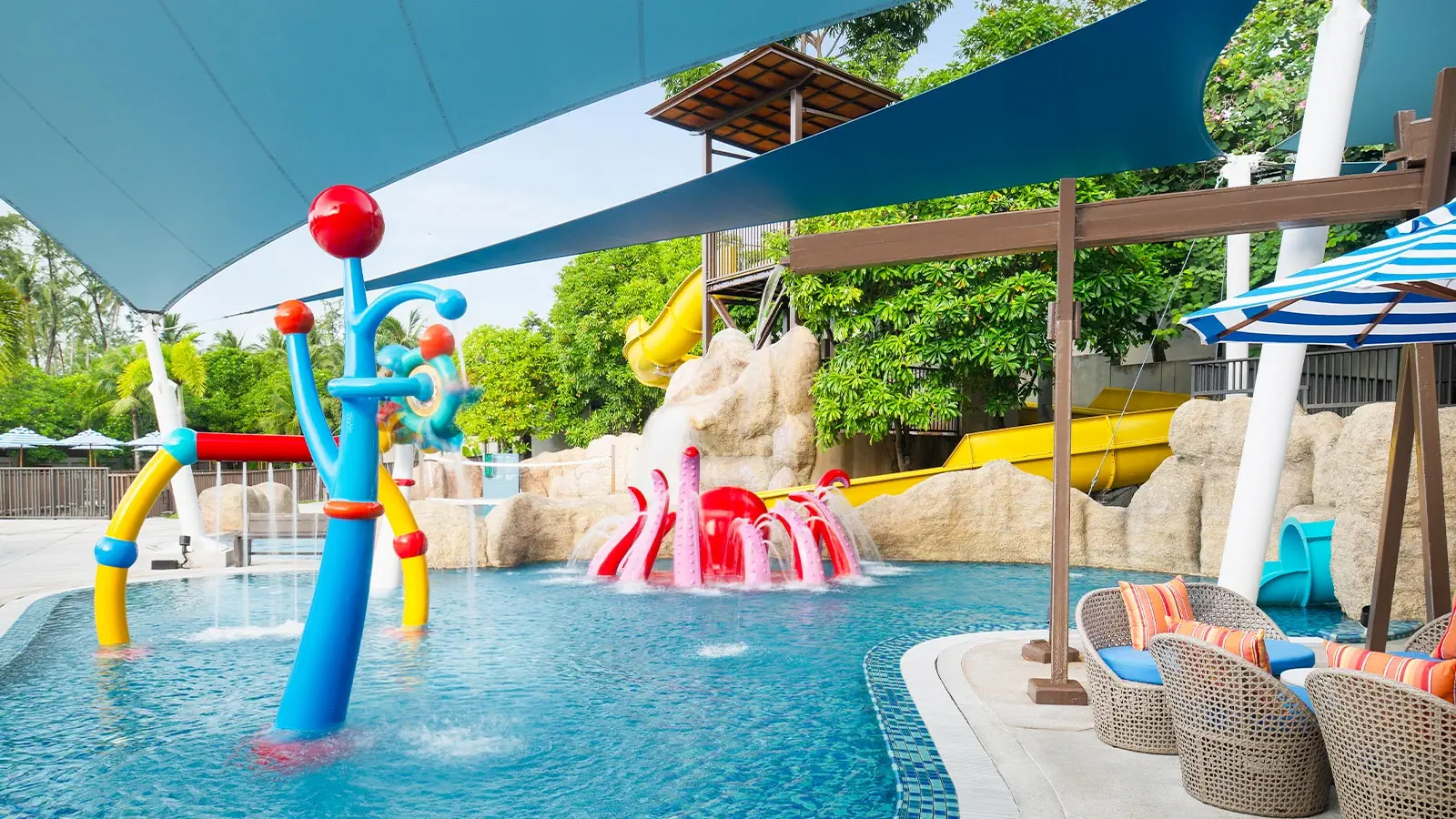 Colorful children's pool at Ozo Phuket with blue and red water play structures, slides, and shaded seating areas surrounded by lush greenery.