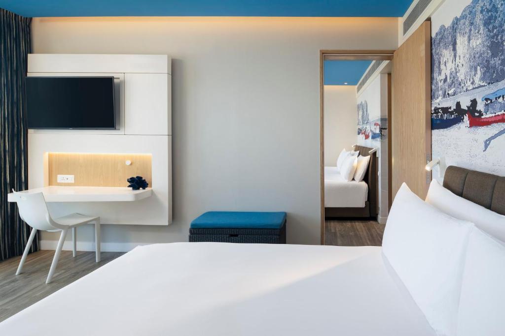 Ozo Phuket deluxe family two-bedroom suite interior with queen bed, wall-mounted TV, desk, and second bedroom visible through open doorway.