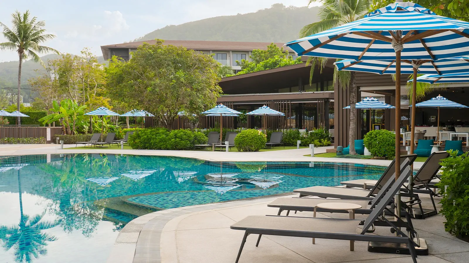 Ozo Phuket resort pool area with turquoise water, blue striped umbrellas, lounge chairs, and modern pavilion surrounded by tropical vegetation and mountains.