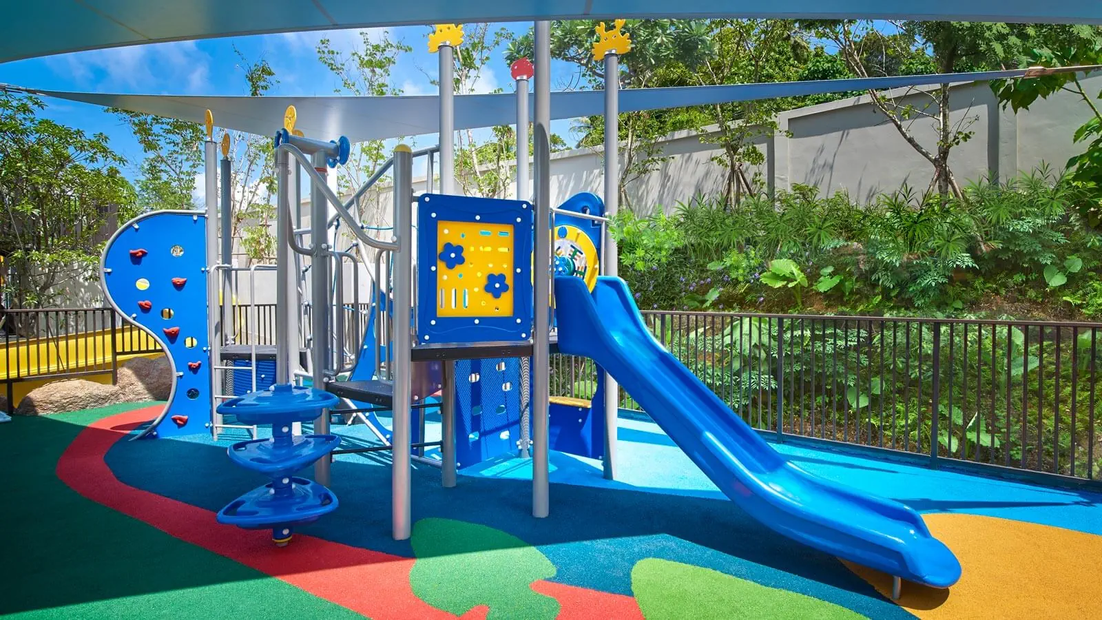 Colorful shaded playground with blue slide, climbing wall, and activity panels on rubber safety surface in Phuket.