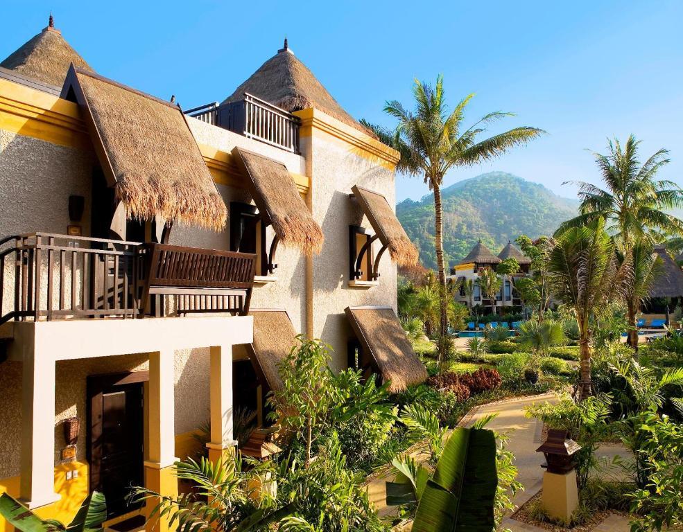 Paradox Karon Phuket resort exterior with thatched-roof bungalows, white walls, yellow trim, palm trees, and mountain backdrop.