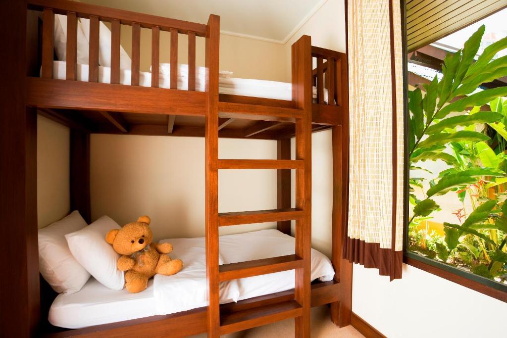 Wooden bunk beds with white bedding and teddy bear in bright villa suite, tropical plants visible through window.