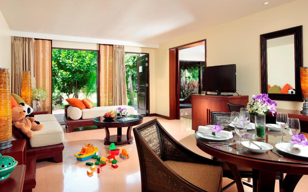 Spacious villa living room with cream walls, dining table with place settings, cream sofa, TV, and garden views through glass doors.