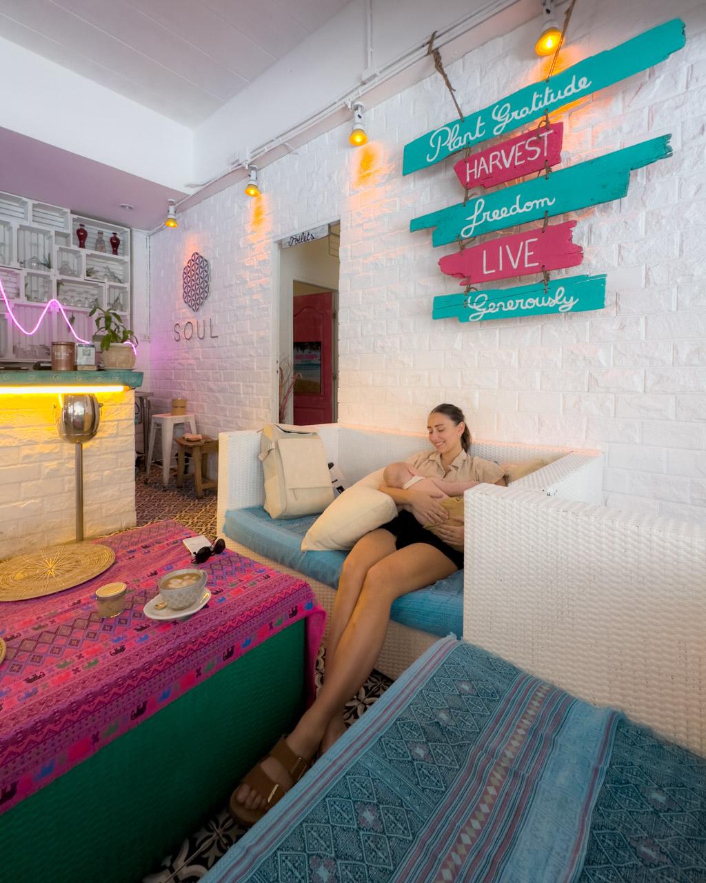 Woman relaxing on bed in colorful cafe with turquoise and pink motivational wall signs, neon lighting, and coffee table.