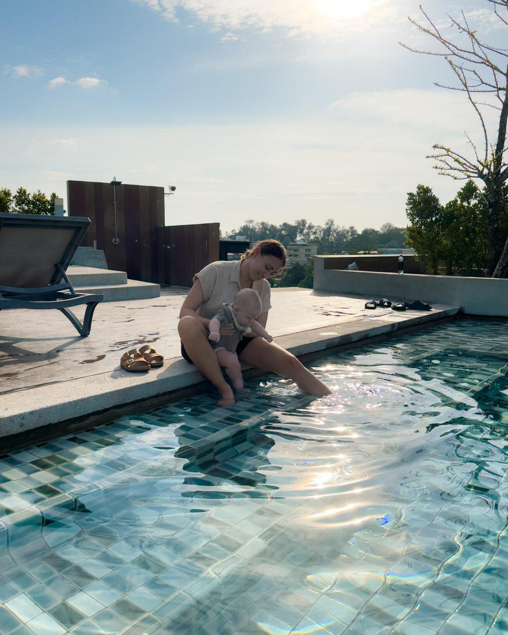 Woman holding infant at edge of rooftop pool in Phuket, dipping baby's feet in water under clear sky.