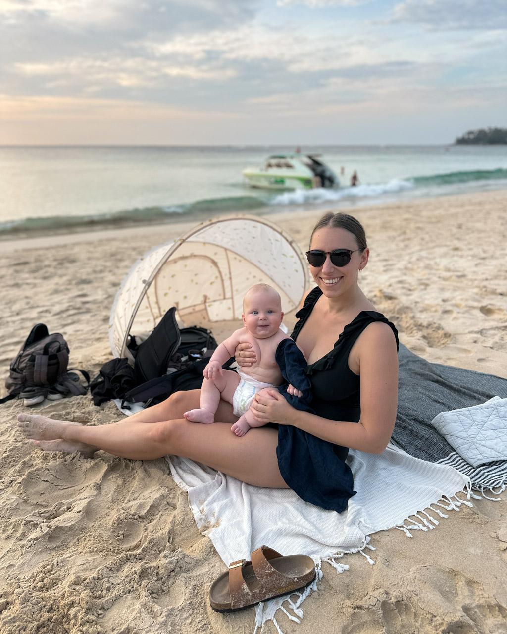Woman in sunglasses holding infant on Phuket beach with pop-up tent and speedboat in background.