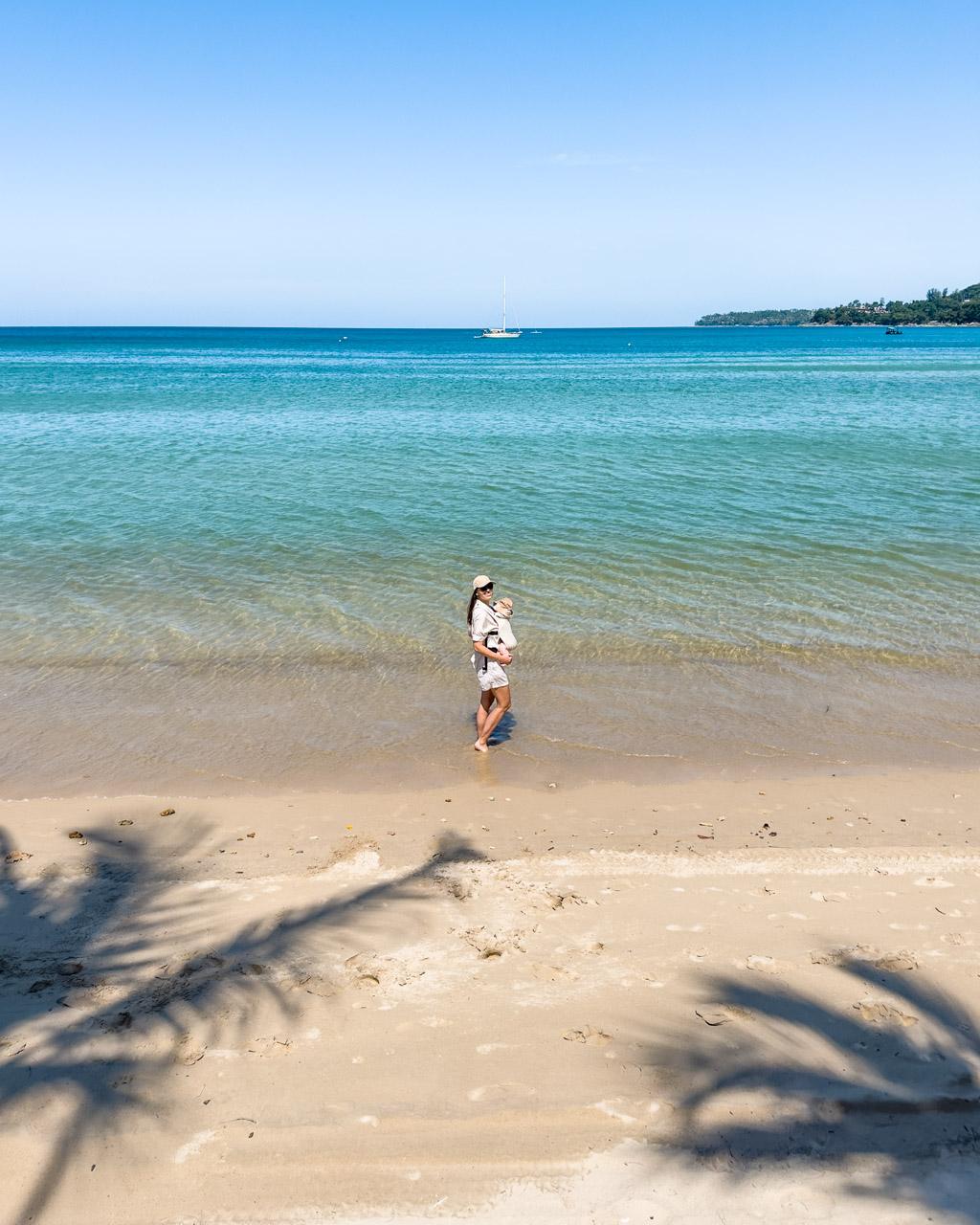 Woman holding child wading in turquoise ocean water at Phuket beach with sailboat and palm trees visible