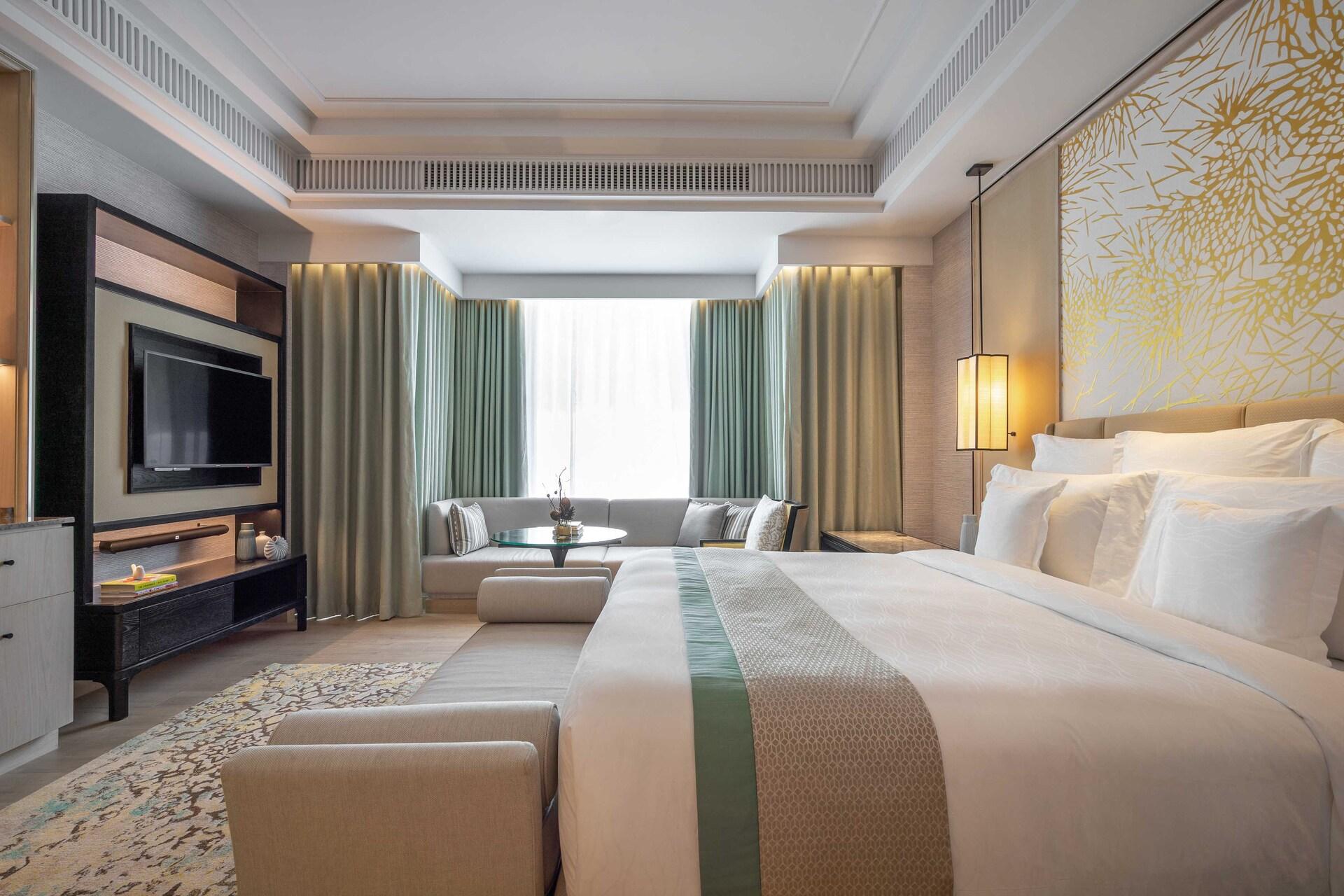 Luxury bedroom with king bed, sitting area with gray sofa, flat-screen TV, floor-to-ceiling curtains, and gold geometric wallpaper.