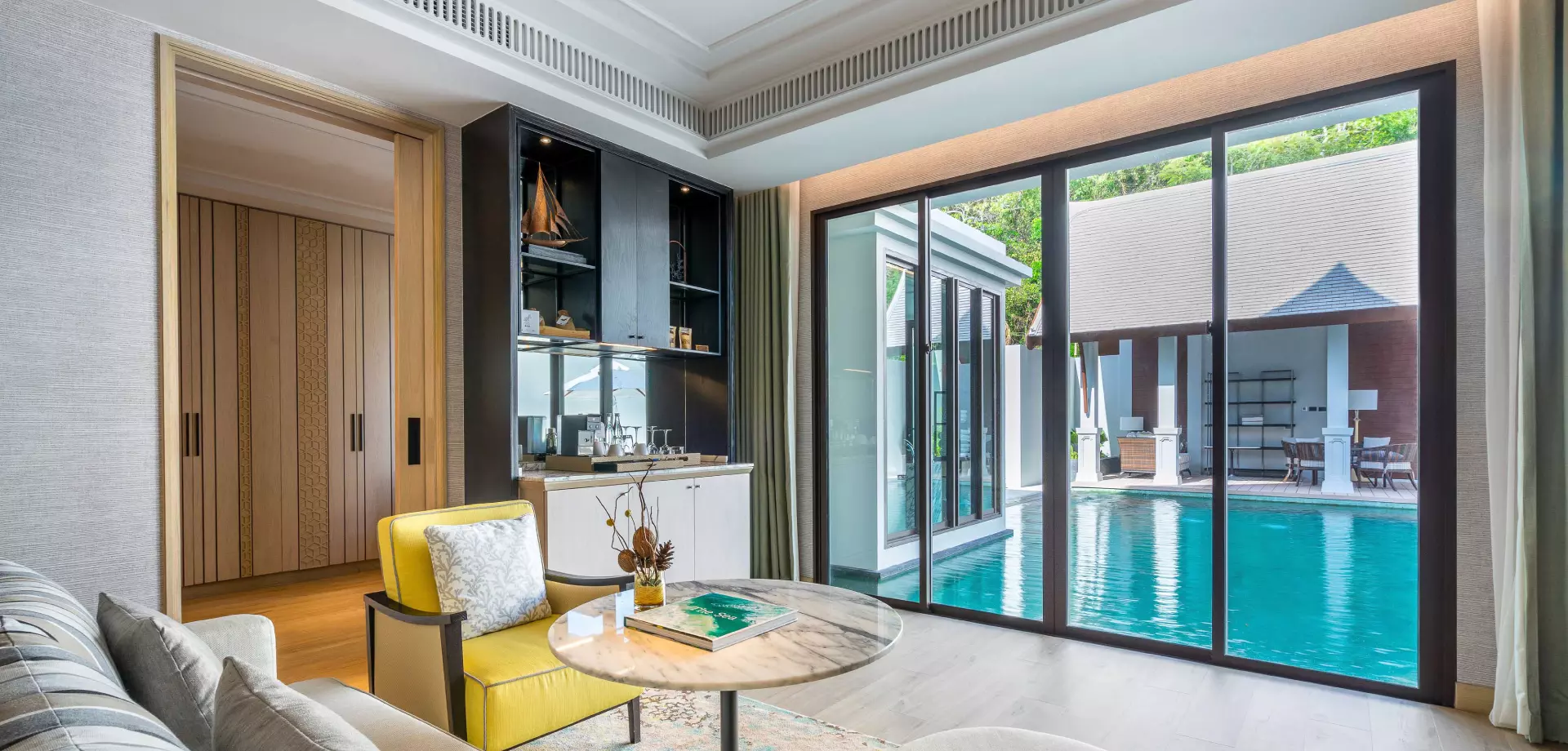 Luxury villa interior with yellow accent chair, marble table, black bar cabinet, and floor-to-ceiling windows overlooking turquoise swimming pool.