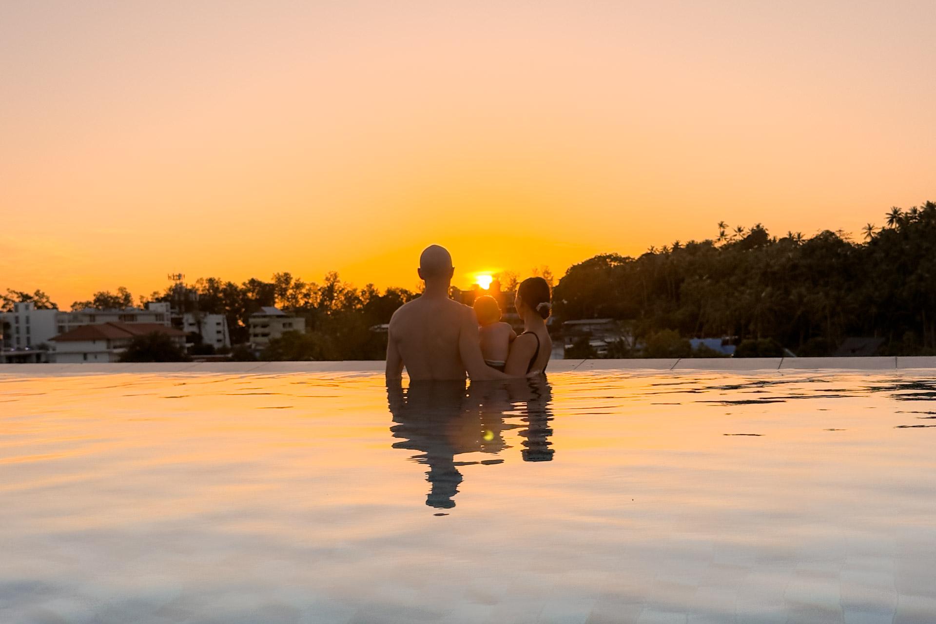 Father and two children swimming in infinity pool at sunset overlooking Phuket tropical landscape with palm trees.