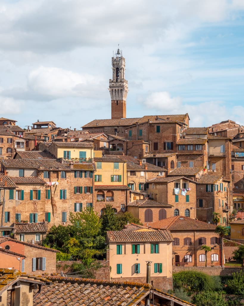 Top of Torre del Mangia behind houses on hill in Siena