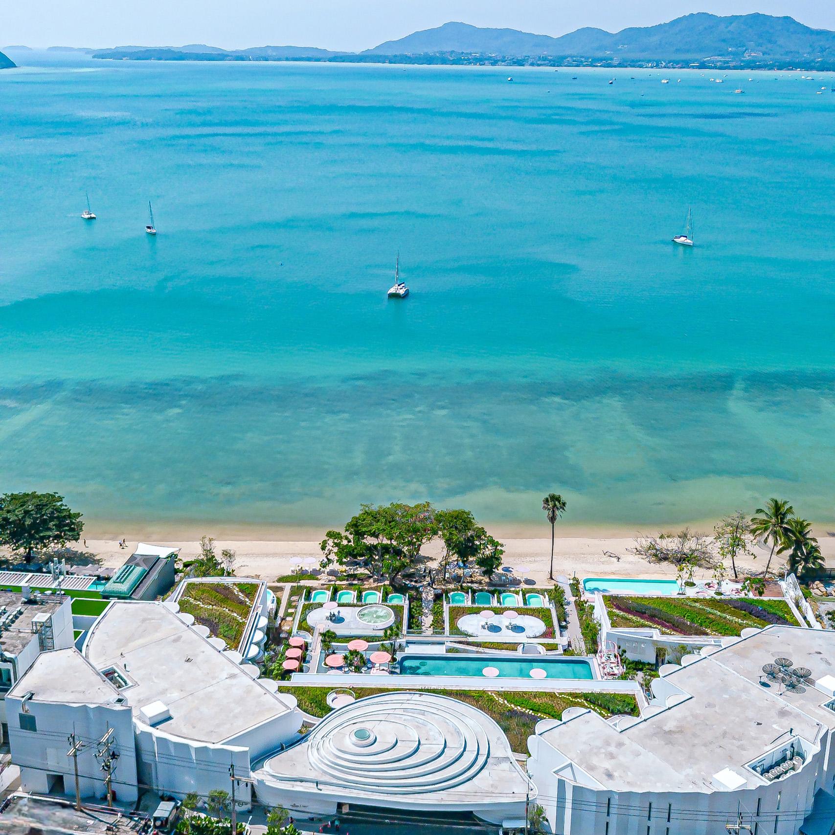 Aerial view of Veranda Resort Phuket with multiple pools, manicured gardens, sandy beach, turquoise waters, and moored sailboats with mountain backdrop.