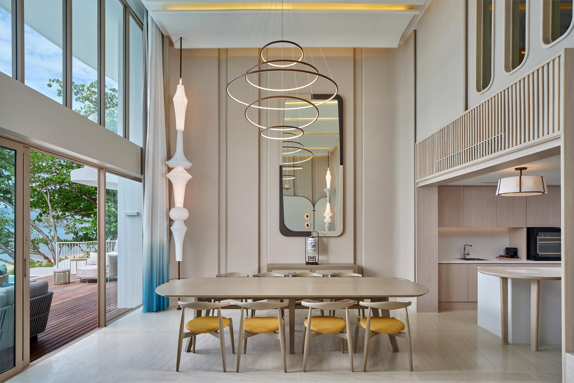 Three-bedroom villa living room with modern dining table, yellow chairs, geometric pendant lights, and floor-to-ceiling windows opening to ocean veranda.